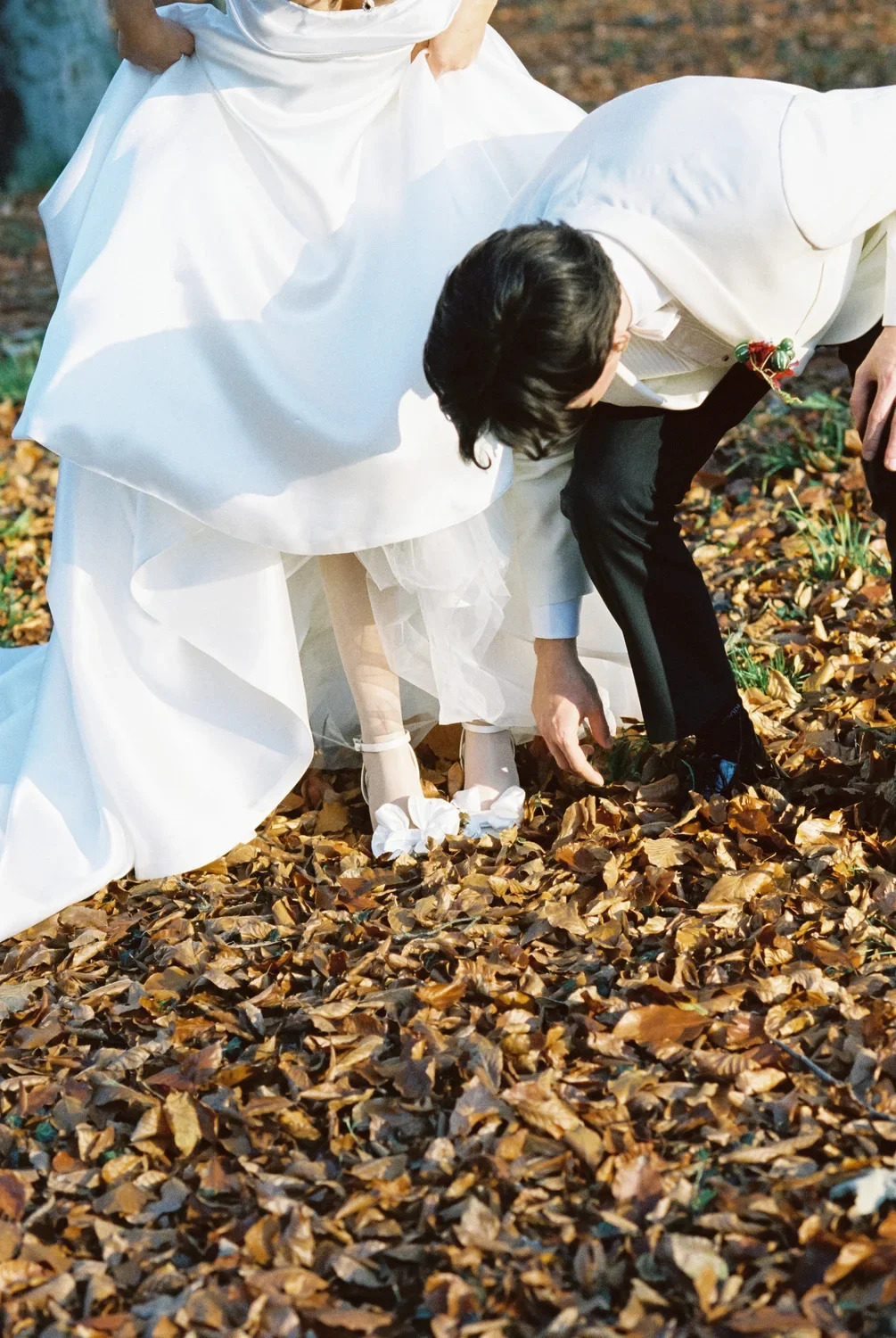Close-up of groom helping the bride with her shoe in fallen autumn leaves, authentic and unposed documentary moment.