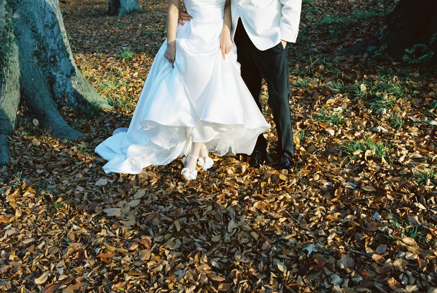 Artistic close-up focusing on the bottom of a wedding dress and shoes amidst deep autumn leaves on the ground.