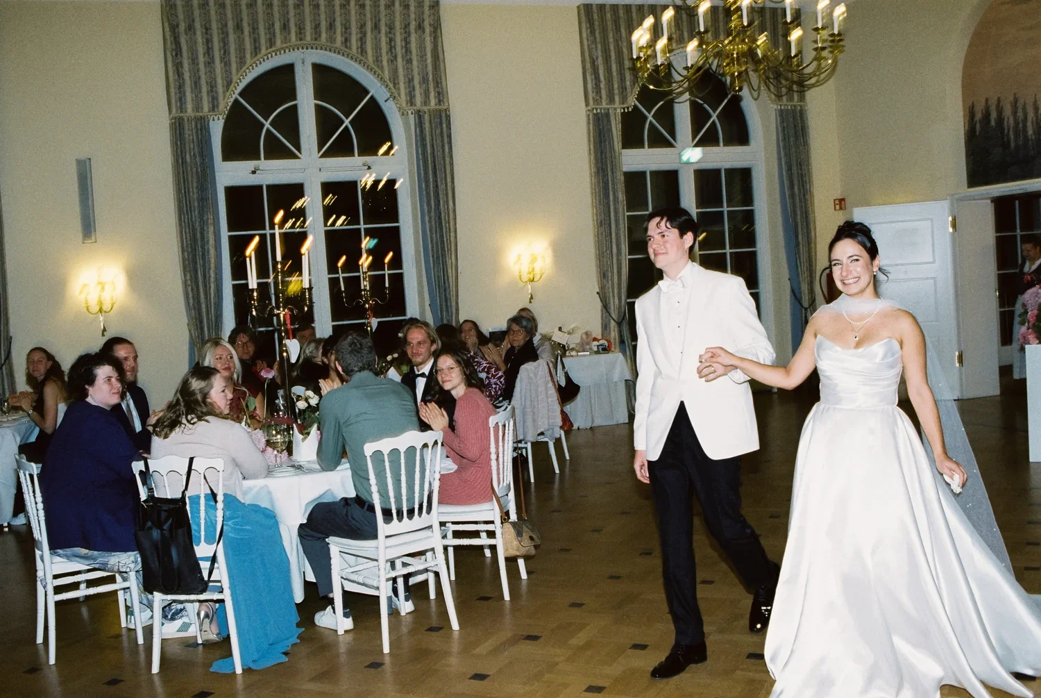 The couple walks past dinner tables, capturing the candid reactions of guests under warm hall lighting.