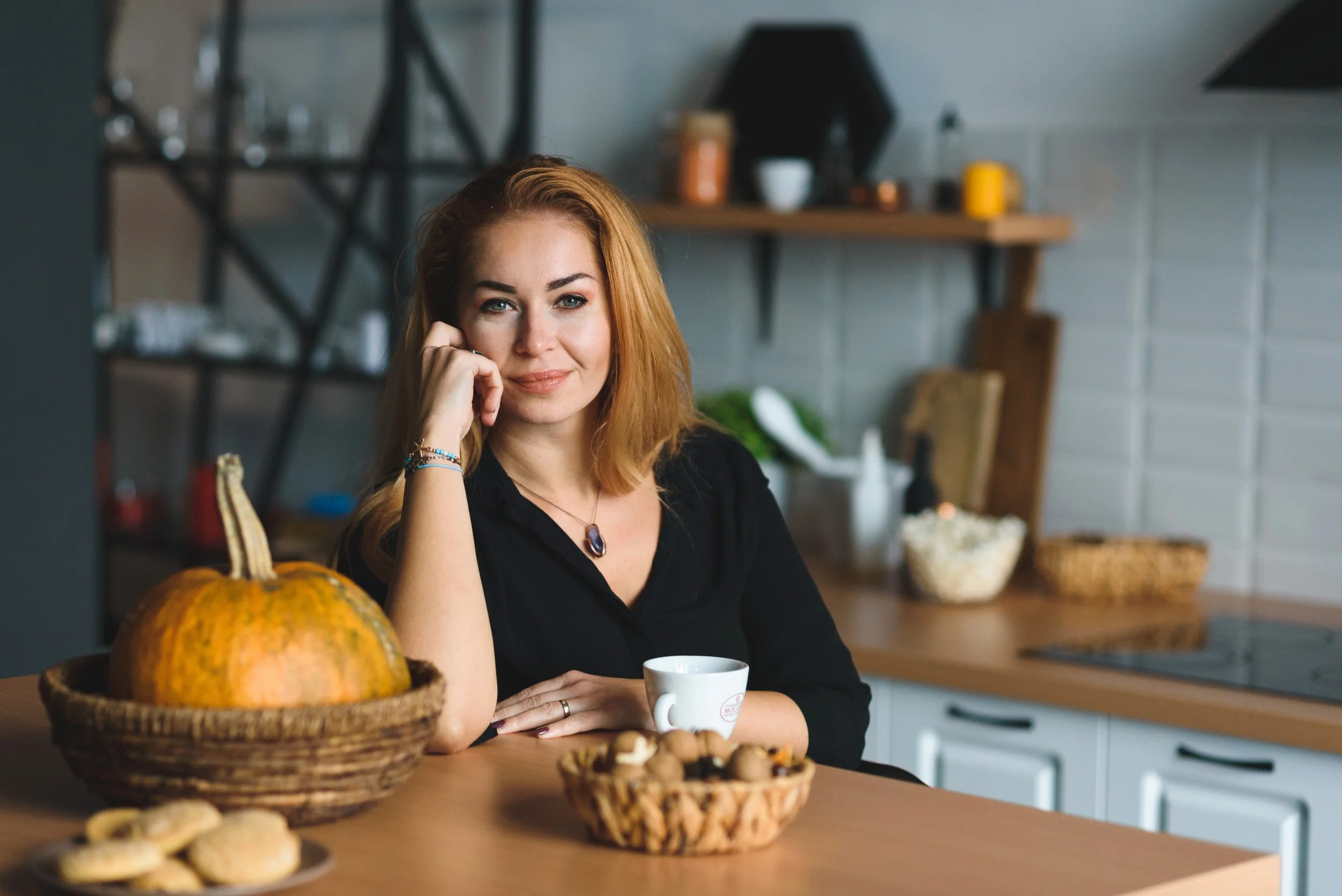 Eine Frau mit roten Haaren sitzt an einem Küchen Insel, umgeben von Keksen, einer Kanne, einer Tasse und einem Kürbis in einem rustikalen Küchenraum.