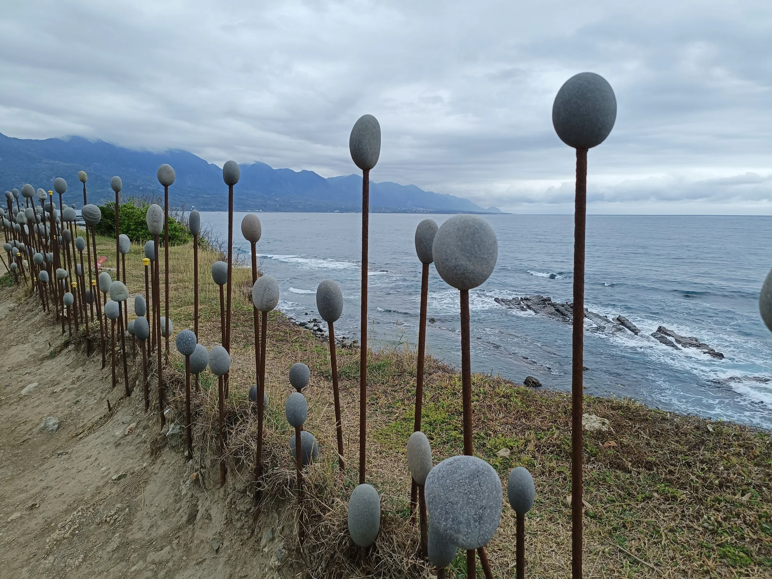 Art installation of stones on metal rods by the sea, with mountains and overcast sky in the background in Taiwan.