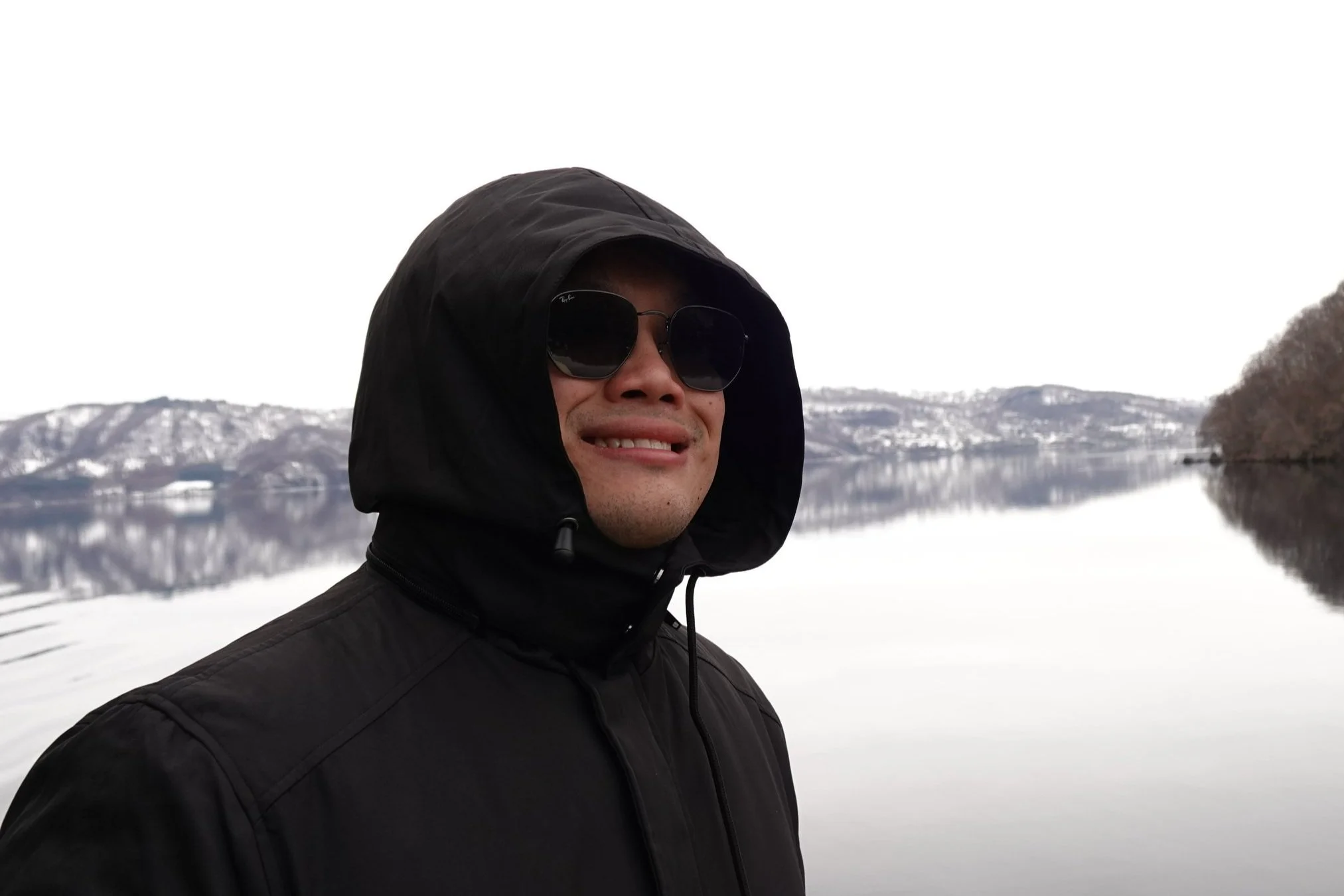 A man wearing sunglasses and a black jacket with a hood stands outdoors near a calm body of water with snow-covered mountains in the background.