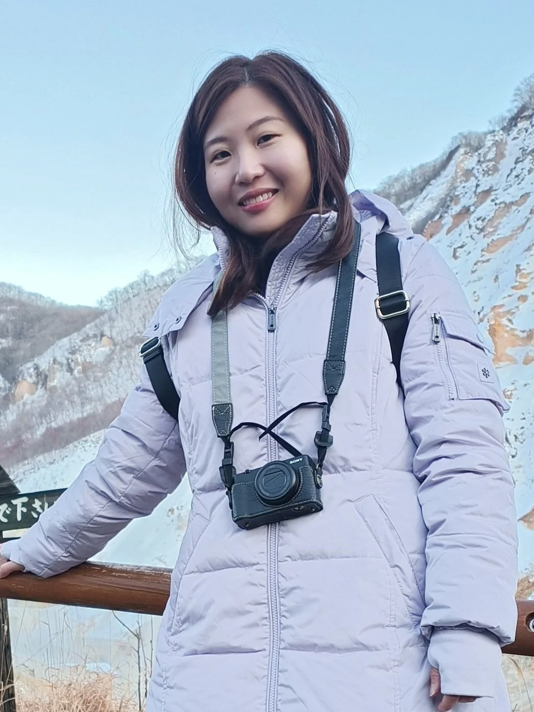 A young woman in a light purple winter jacket with a Sony camera hanging around her neck, standing outdoors on a wooden railing against a snowy mountain landscape.