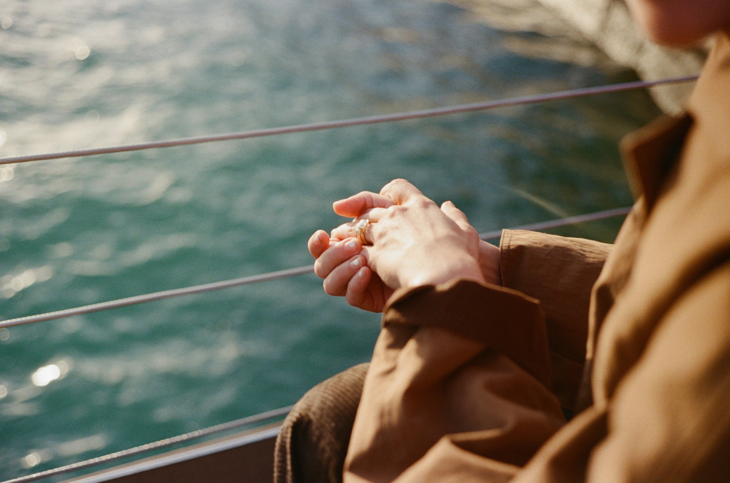 Person sitting on a boat, hands clasped, near a body of water, with a railing and ropes visible.