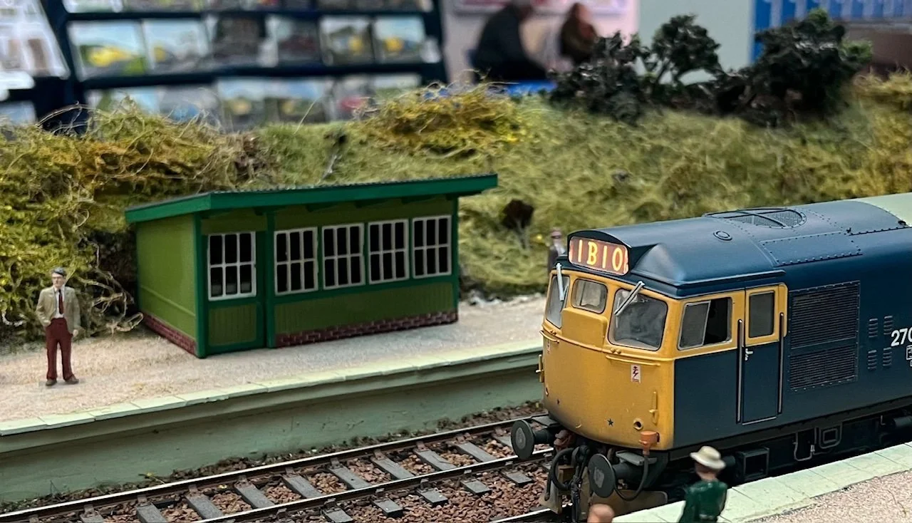 A model train station scene featuring a yellow and blue train with the number 2710, a small green station building with multiple windows, and a miniature figure of a man in a suit and hat near the tracks. There are also some moss and other greenery i