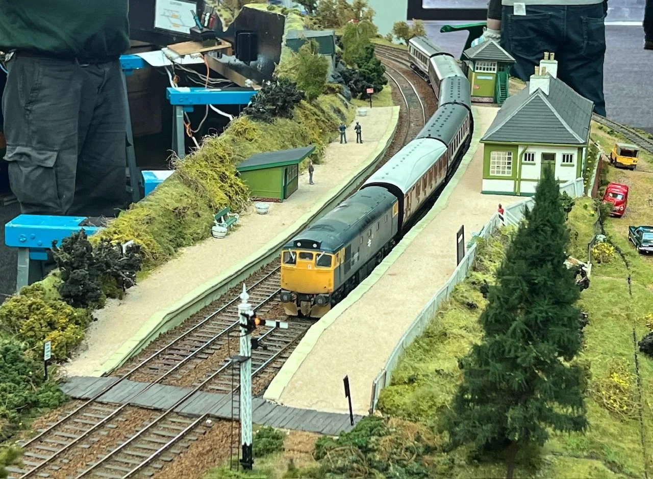 Model train layout with a black and yellow train on tracks, surrounded by miniature trees, houses, and figures, with control equipment visible in the background.