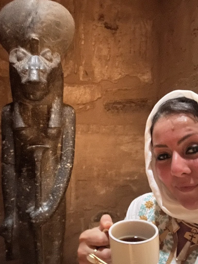 A woman wearing a white headscarf holding a white mug of coffee takes a selfie next to an ancient Egyptian statue of a man with a headdress, set against a sandstone wall.