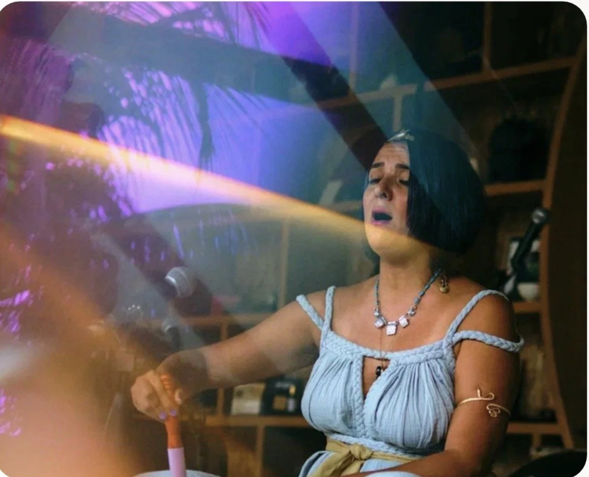 A woman with black hair and a blue dress sings passionately into a microphone during a performance or karaoke, with colorful stage lighting reflecting around her.