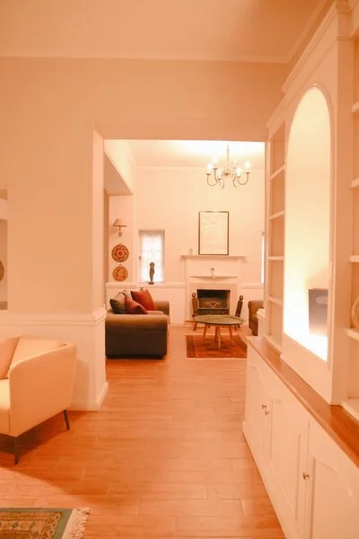 Living room with white walls, a gray couch with orange and beige pillows, a white armchair, hardwood floor, and a fireplace with a mirror above, chandelier hanging from ceiling.