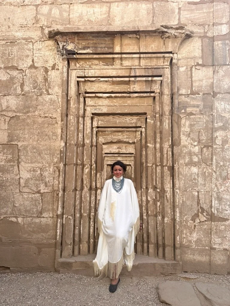 A woman standing in front of an ancient, weathered stone wall with a carved rectangular doorway behind her.