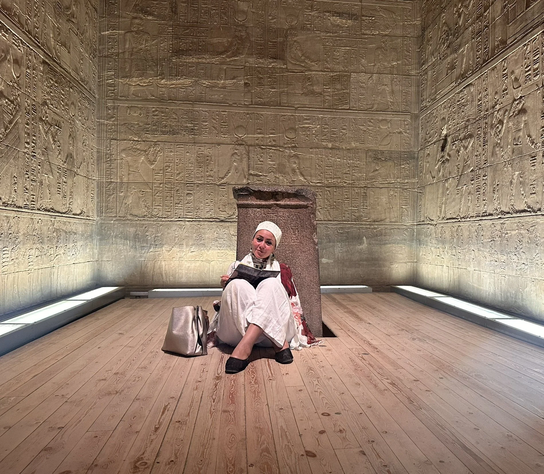 A woman wearing a white headscarf and dress sits on the wooden floor in front of a wall covered with ancient Egyptian hieroglyphs, holding an open book, with a beige purse beside her.