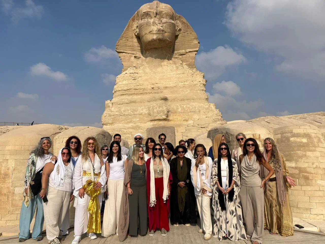 People sitting and standing under a large tree near a rocky landscape and a small stream, with the sun shining through the branches.