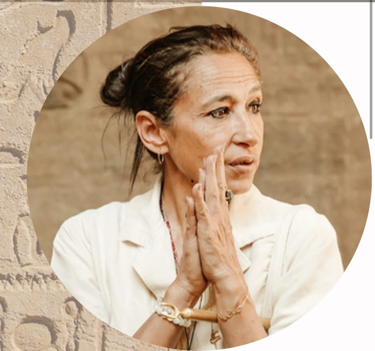 A woman with brown hair tied in a bun, wearing a white jacket, and multiple jewelry pieces, touching her face and looking to the side against a textured wall background.