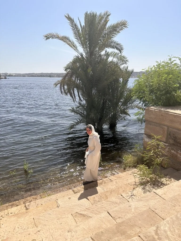 A woman dressed in white standing by a body of water with a palm tree in the background under a clear blue sky.