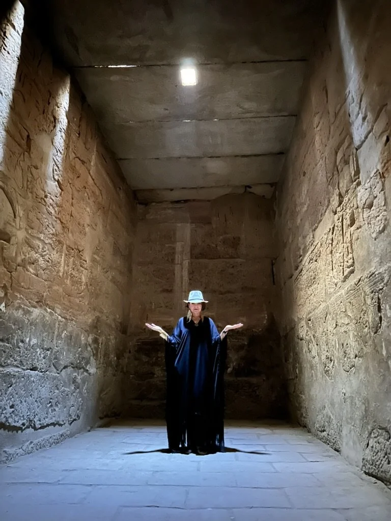 A woman wearing a hat and black gown standing inside an ancient stone structure with her arms open.