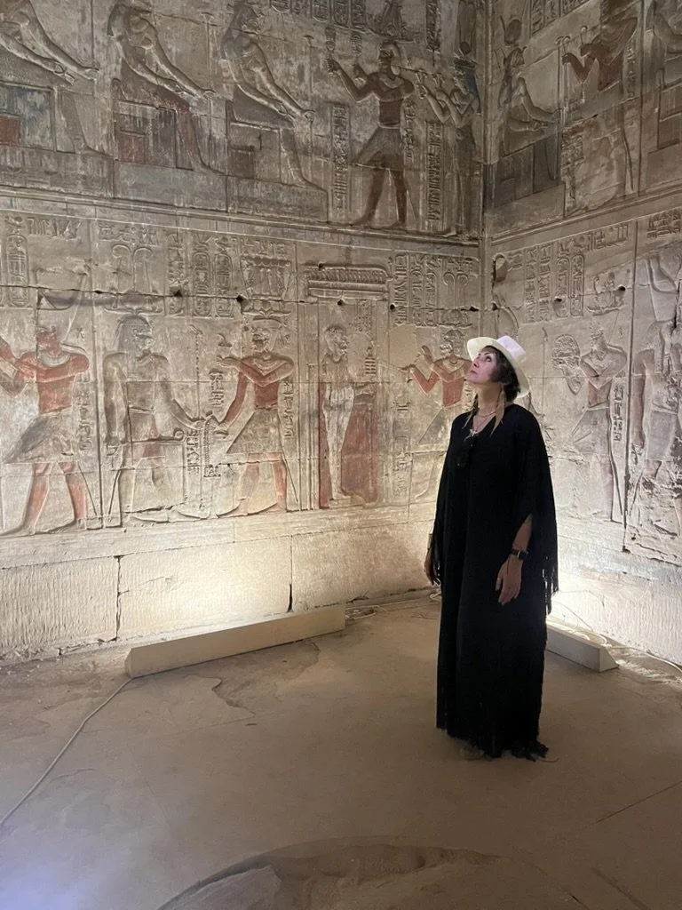 A woman wearing a white sun hat and black dress looking up at ancient Egyptian hieroglyphic and fresco wall carvings in a museum or archaeological site.