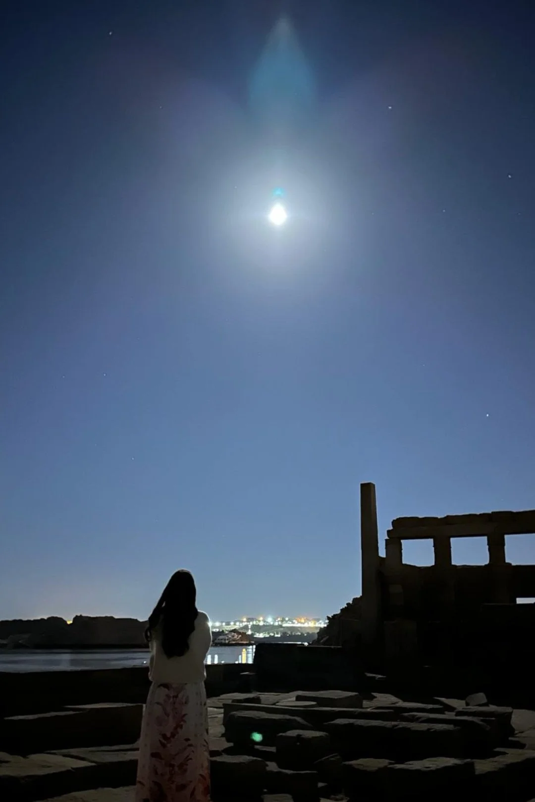 A woman with long hair stands near a body of water at night, looking at a bright full moon in a clear sky with some stars, with distant city lights in the background.