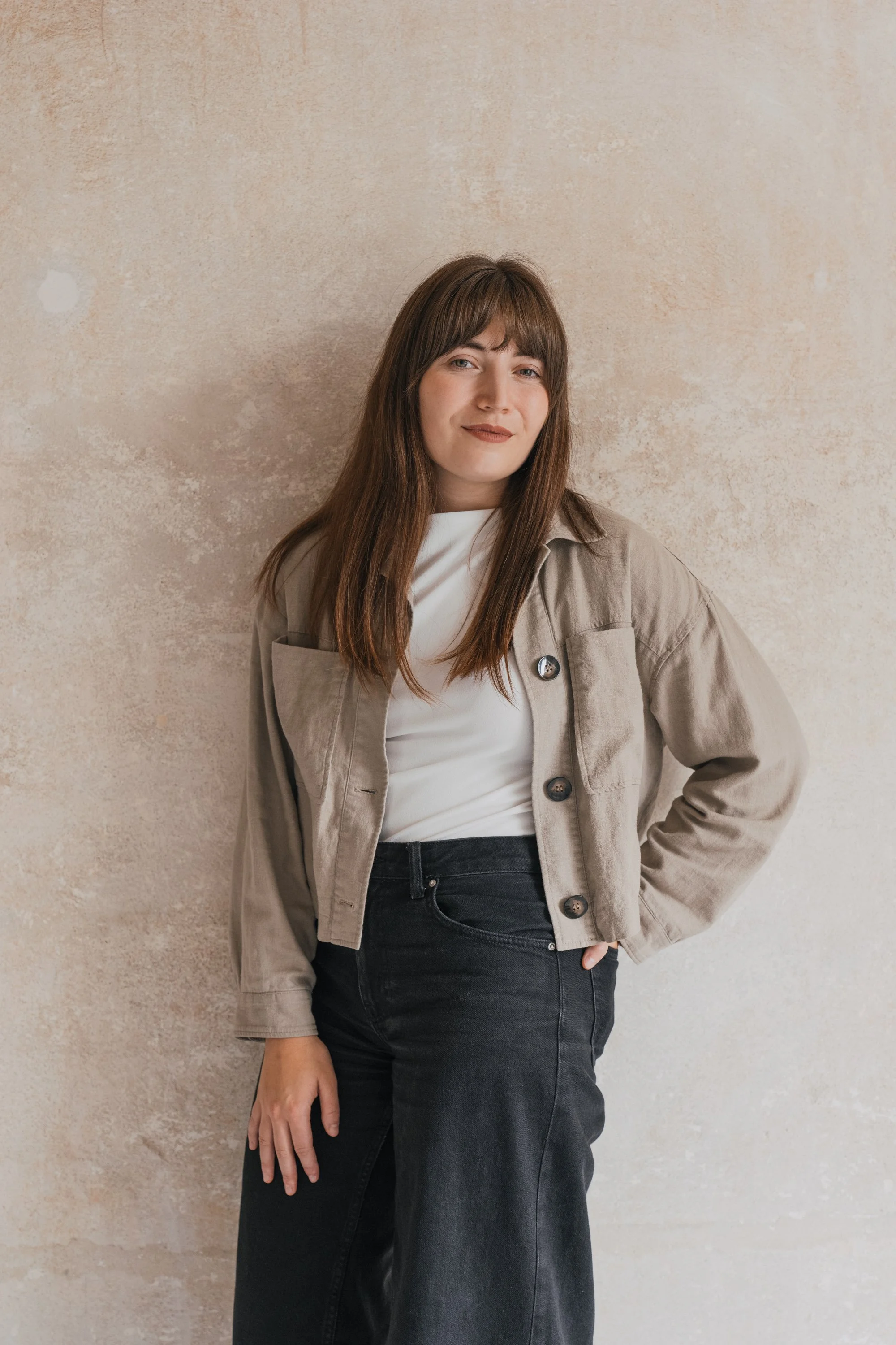 A young woman with long brown hair, wearing a gray sleeveless top, blue jeans, beige socks, and sneakers, sitting on a green stool against a beige textured wall, smiling at the camera.