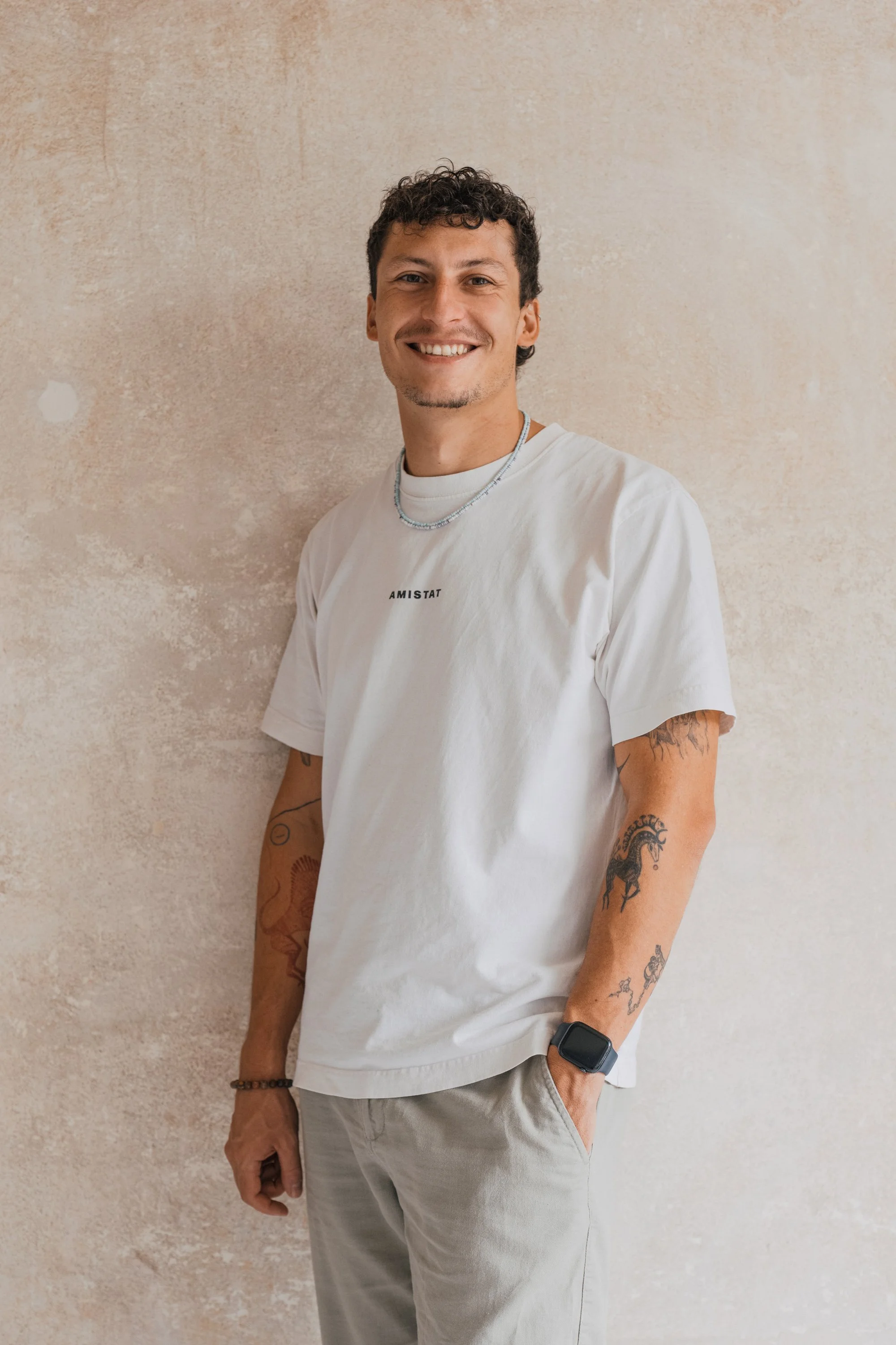 A young man with tattoos, wearing a white t-shirt with 'AMISTAT' on it, jeans, and sneakers, sitting on a turquoise stool against a textured beige wall.