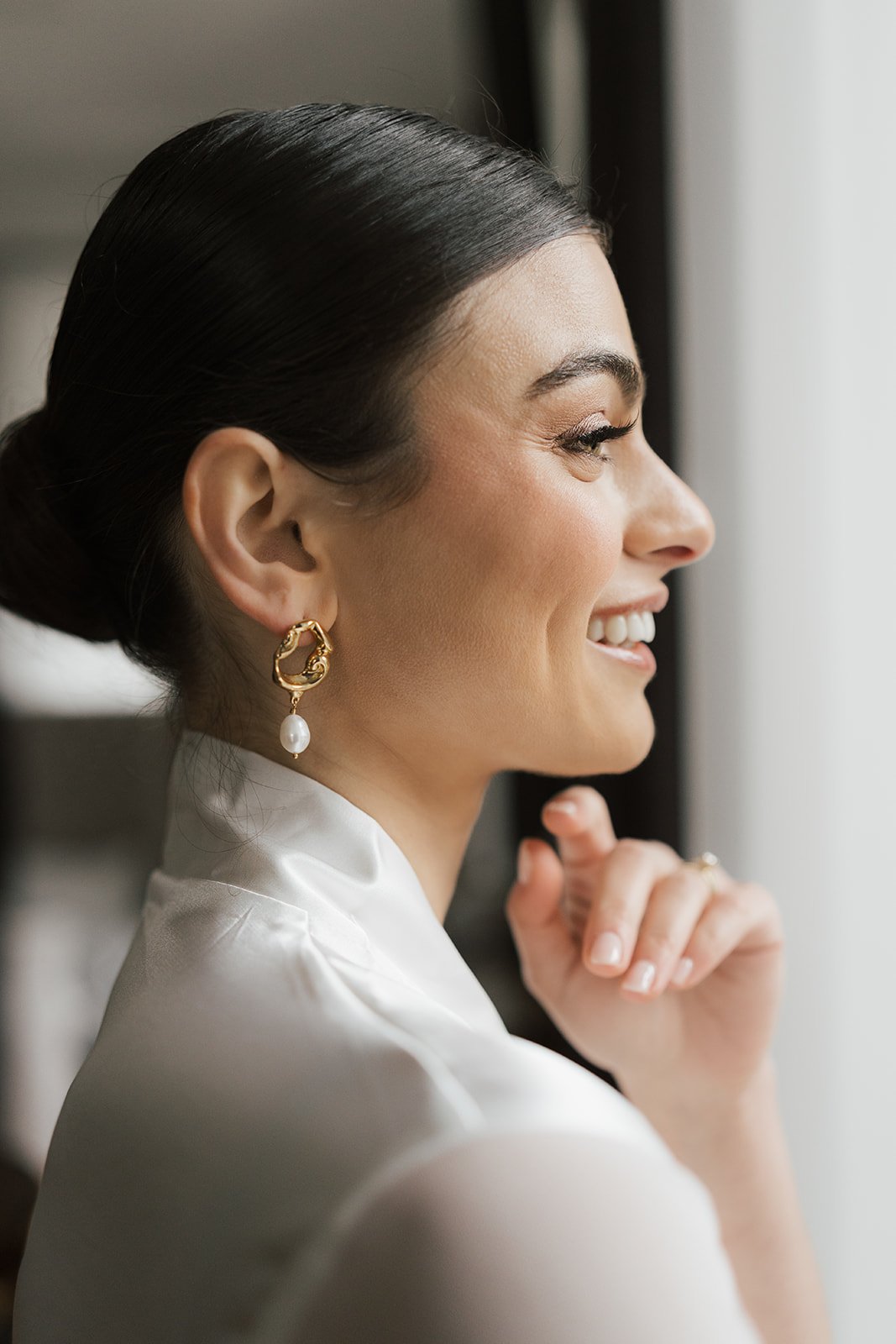 A woman with dark hair styled in a bun, smiling while looking out a window. She is wearing gold hoop earrings with pearls and a white satin robe.