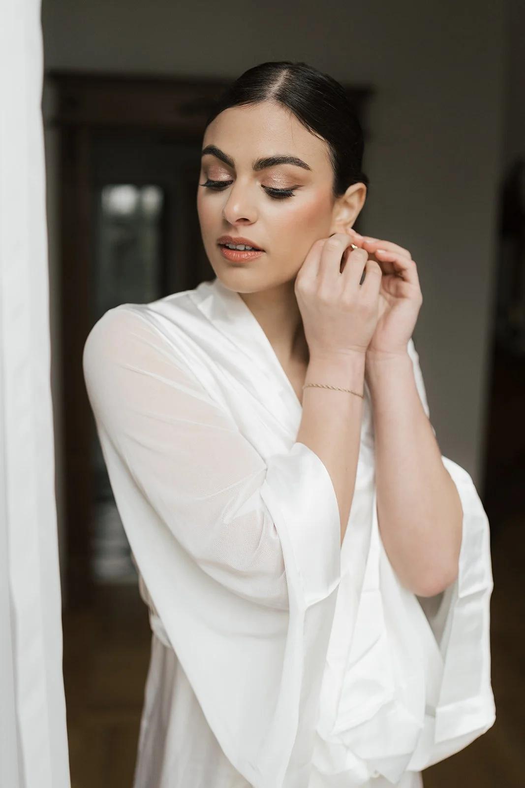 A woman with dark hair and makeup is putting on earrings, wearing a white garment.
