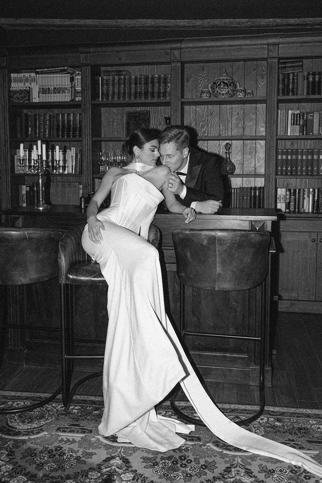 A black and white photo of a woman in a white dress and a man in a tuxedo inside a library or study with wooden bookshelves, a chandelier, and a patterned rug.