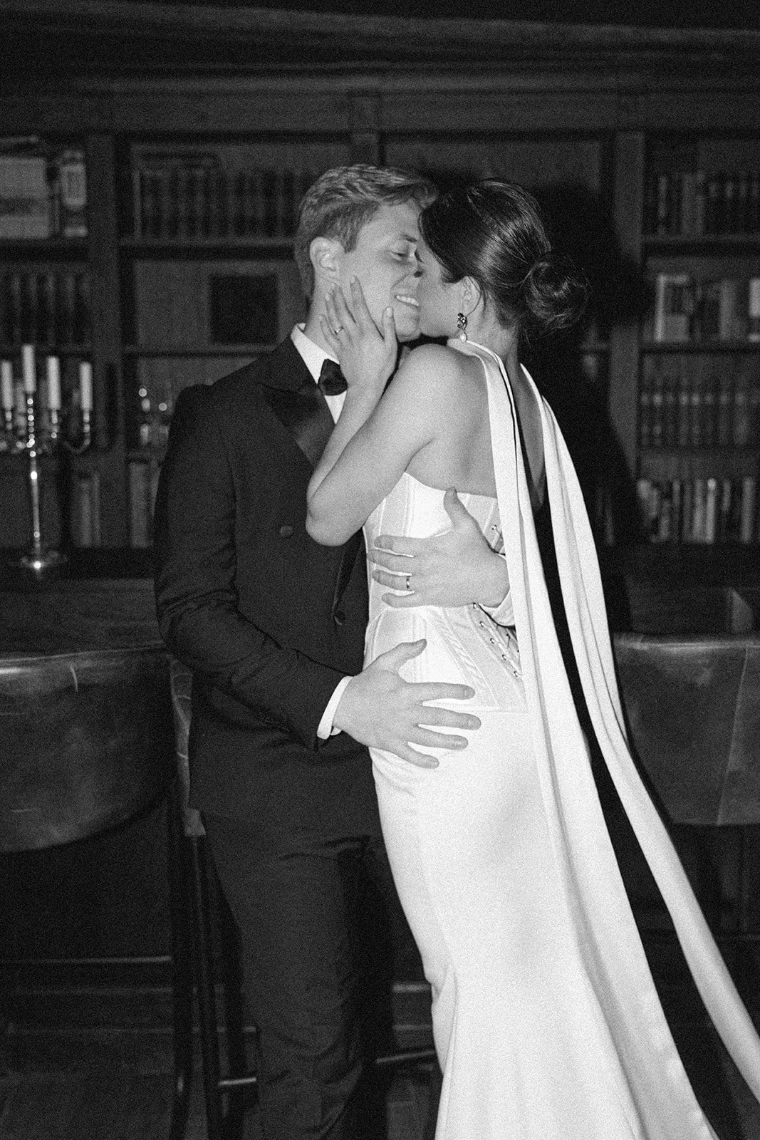 A black and white photograph of a couple sharing a kiss at a formal event, with the man in a tuxedo and the woman in a white gown, in front of a bookshelf and candleholders.