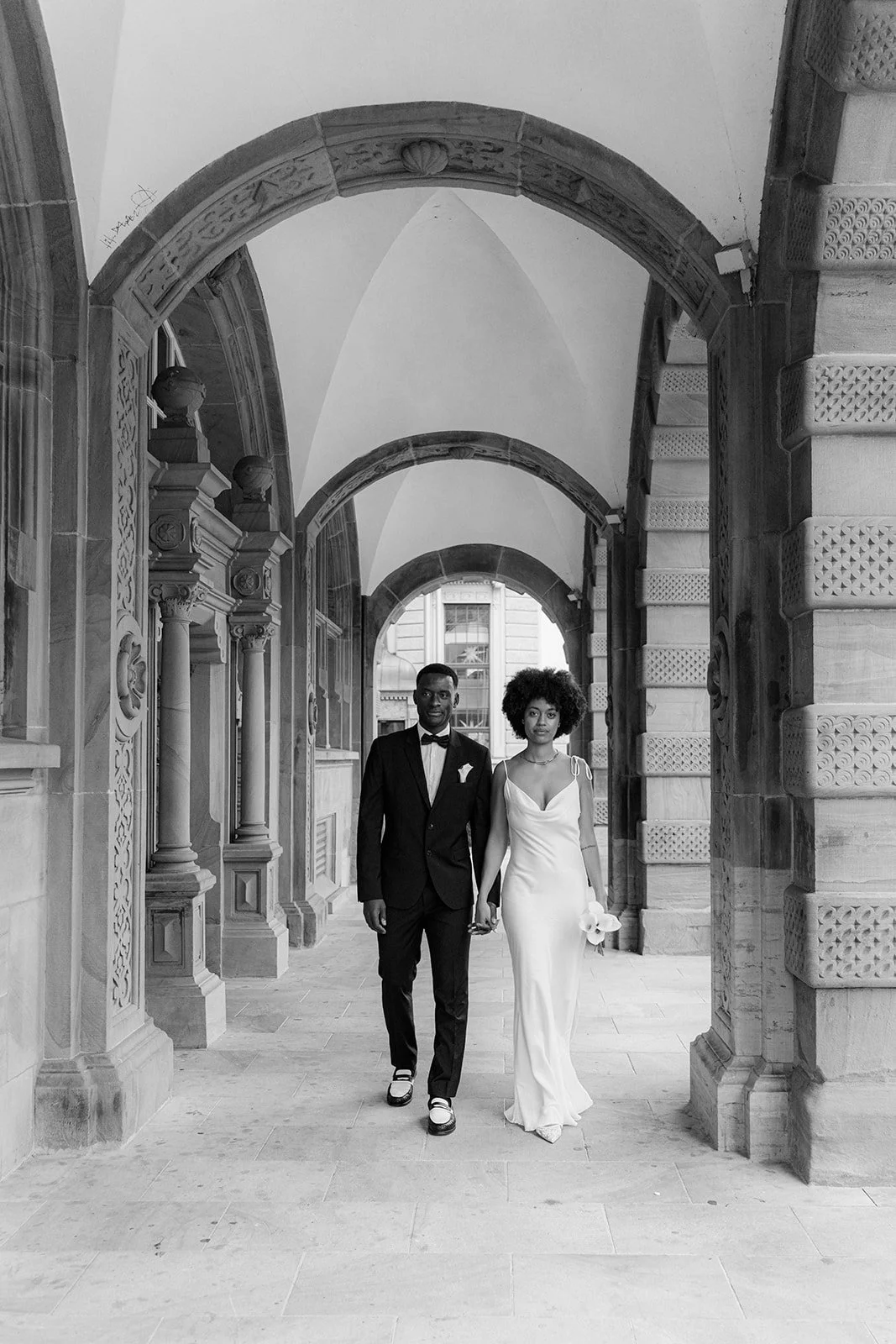 A couple dressed in wedding attire walking through an arched stone passageway, holding hands. The groom wears a tuxedo and the bride wears a white gown holding a flower.