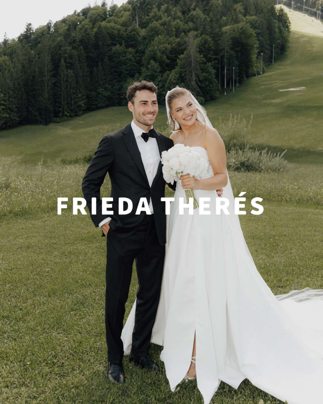 A happy bride and groom standing outdoors on a green field with trees and hills in the background, smiling at the camera. The bride is in a white wedding dress holding a bouquet of white flowers, and the groom is in a black tuxedo with a bow tie.