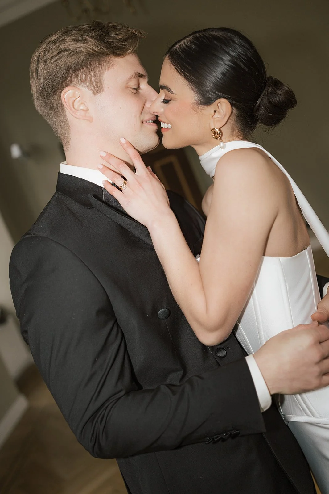 A man and woman sharing an intimate moment, close to kissing. The man is dressed in a black tuxedo, and the woman is wearing a white dress with a high neckline and gold earrings.