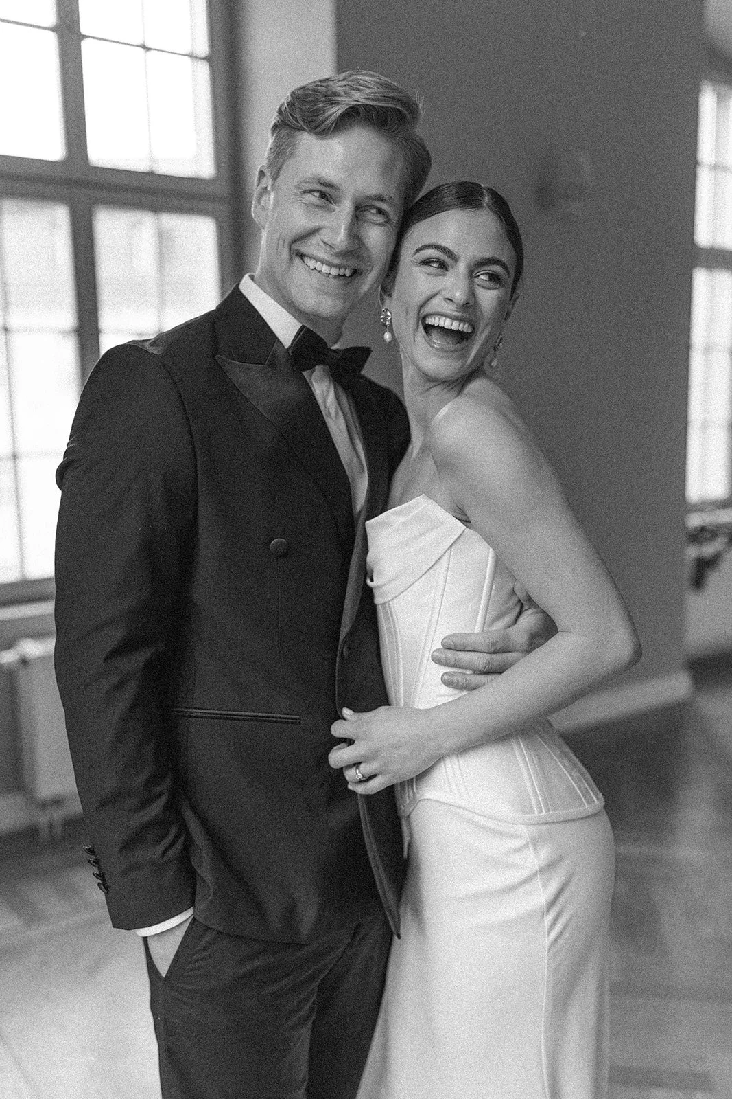 A smiling man and woman in elegant formal attire share a joyful moment indoors with large windows in the background.