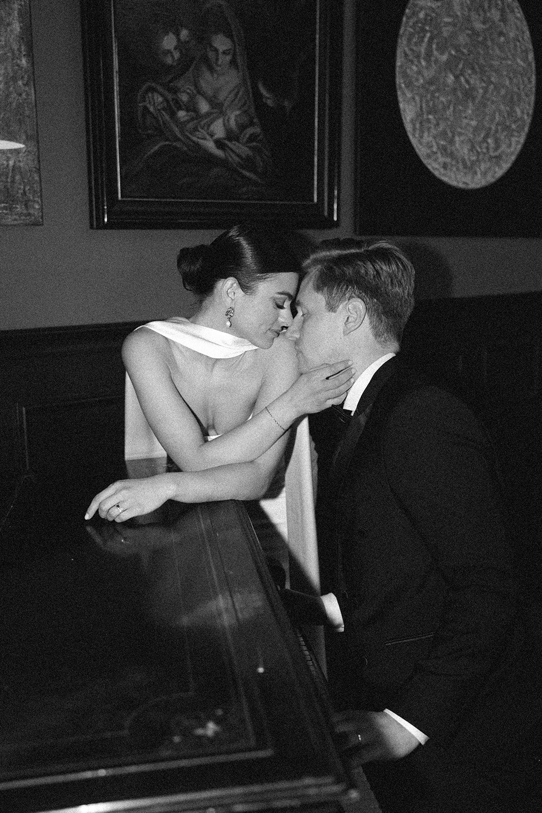 Black and white photo of a couple in an intimate moment, with their foreheads touching and eyes closed. The woman is leaning over a piano, wearing a sleeveless dress and earrings, while the man sits close, dressed in a suit. Artworks are hanging on t