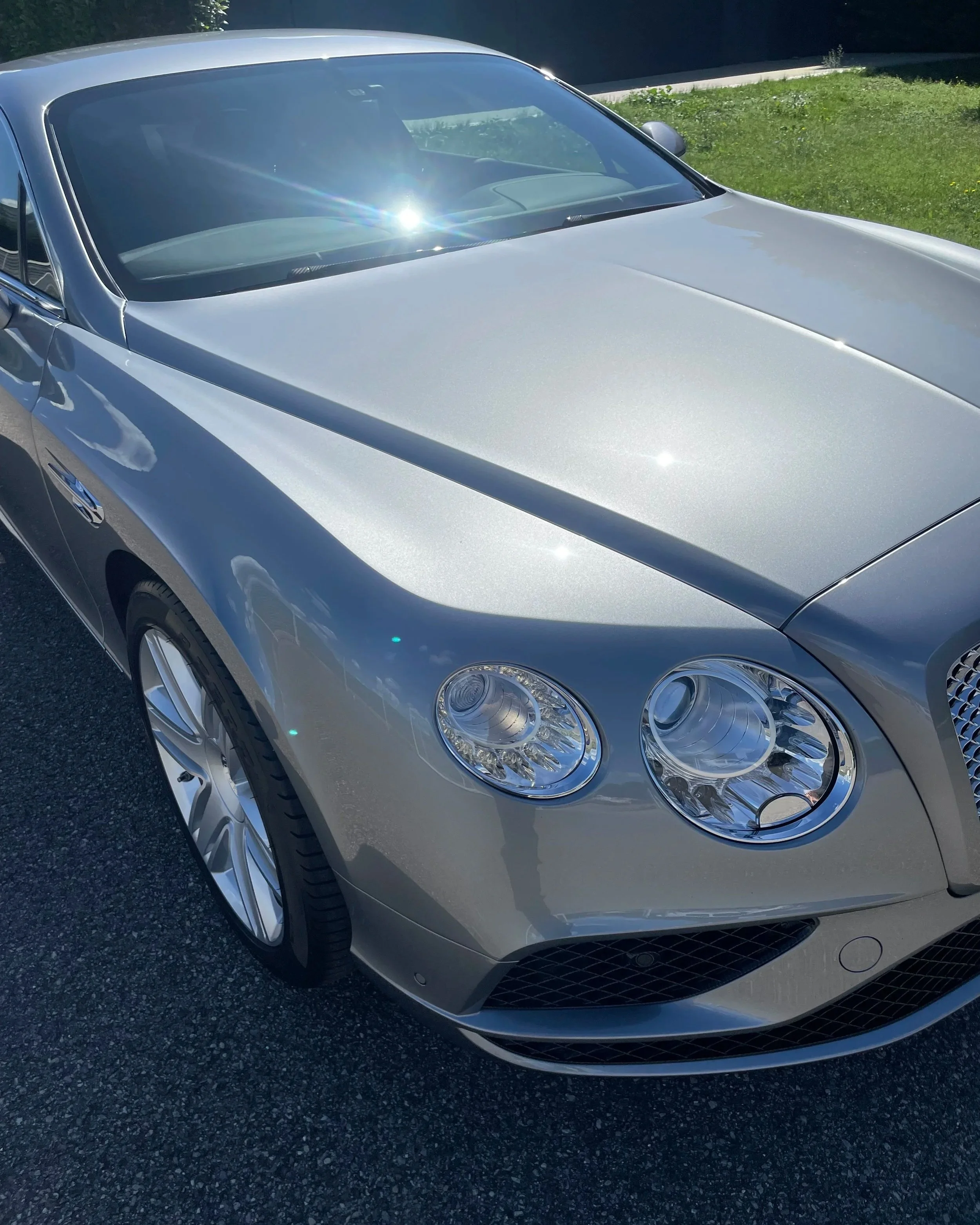 Voiture de luxe grise avec phares ronds et capot ensoleillé sur une route.