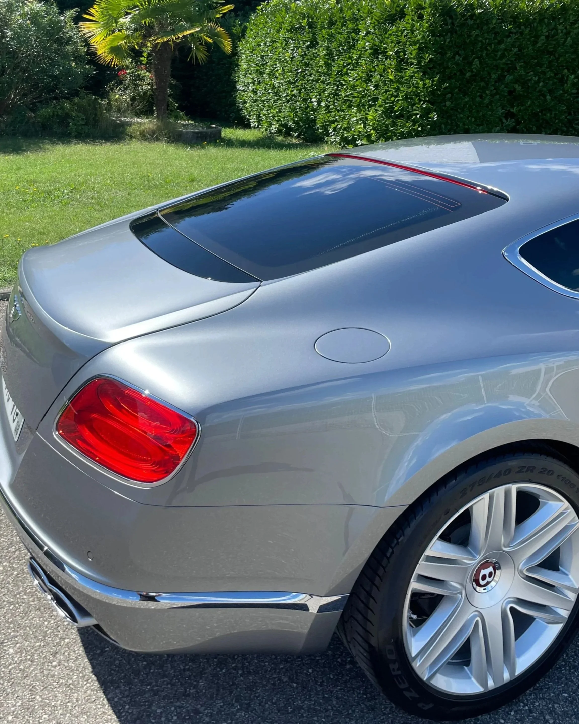 Voiture Bentley silver visible from the back, parked outdoors with green bushes and trees in the background.