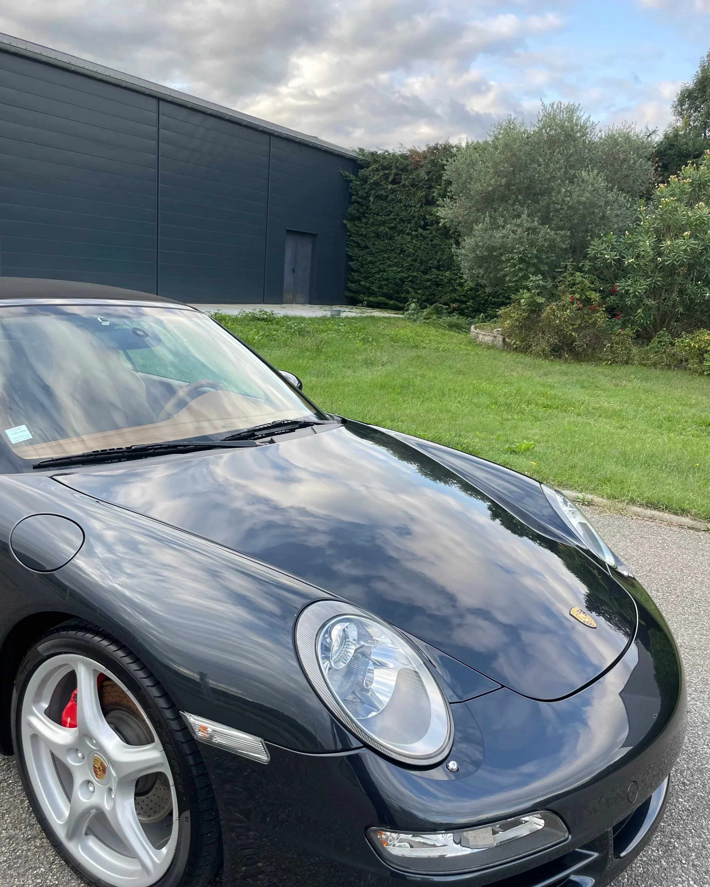 Voiture noire Porsche Carrera dans une allée, avec une maison en arrière-plan, ciel nuageux, et quartiers verdoyants.