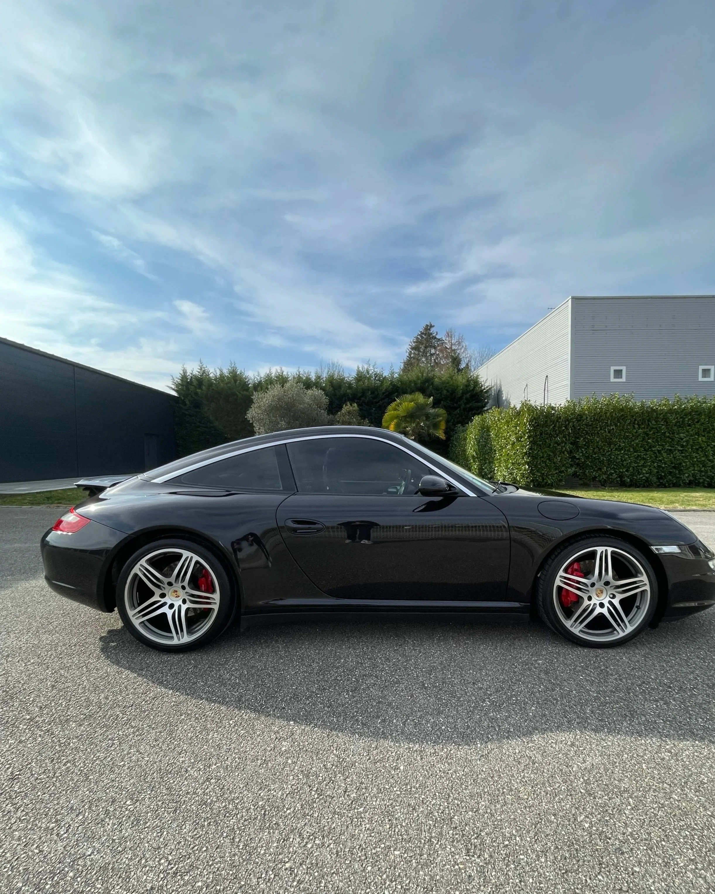 Voiture de sport Porsche noire stationnée sur la route avec un ciel bleu et des bâtiments modernes en arrière-plan.