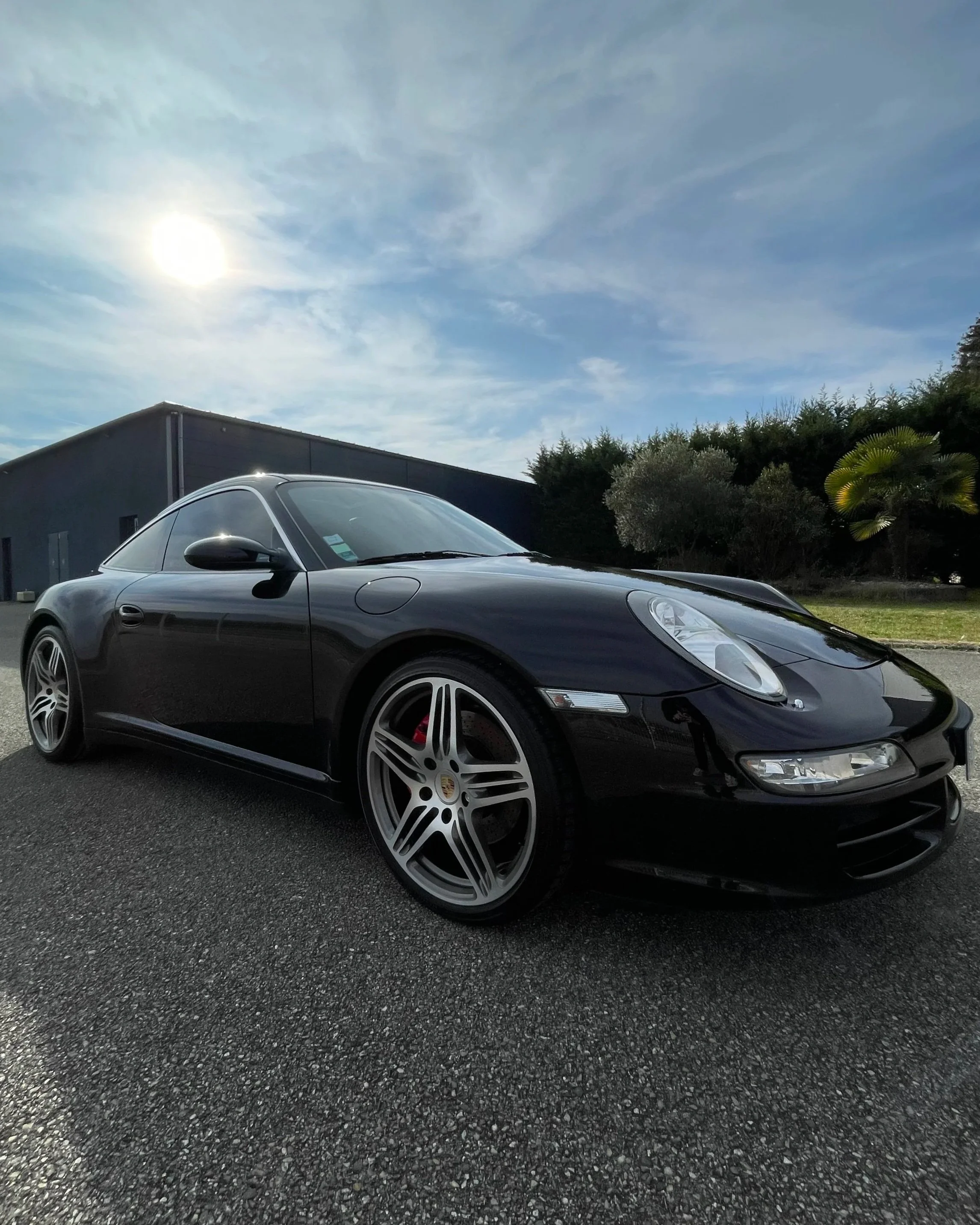 Voiture Porsche noire garée sur une route avec un ciel en partie nuageux et le soleil brillant, entourée de verdure et d'une bâtisse moderne en arrière-plan.