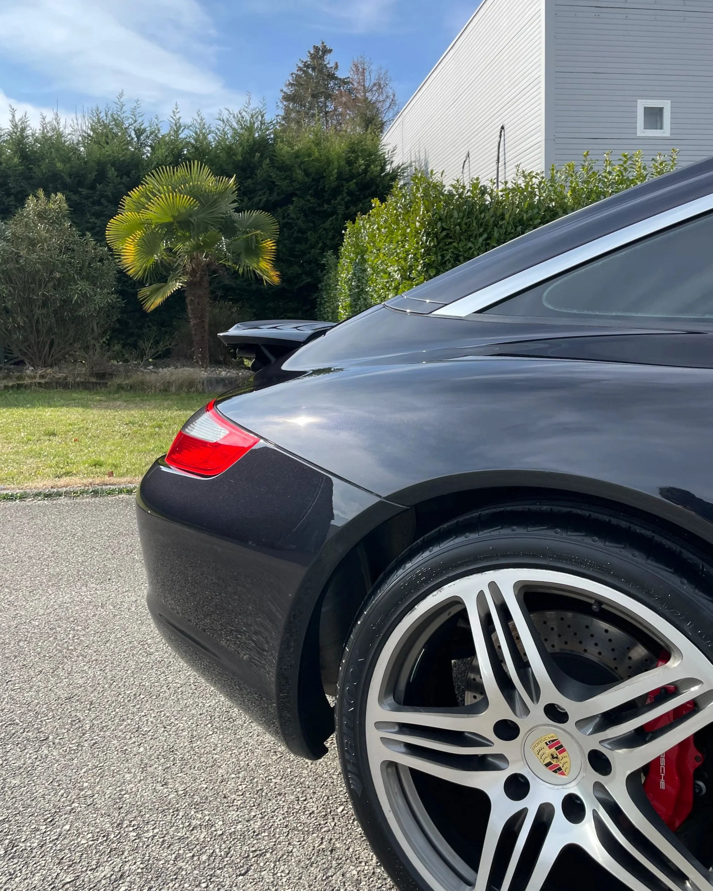 Partie arrière d'une voiture Porsche noire électrique, montrant la roue avec un logo Porsche, la jante, le frein à disque et la calandre rouge, avec un paysage de jardin en arrière-plan.