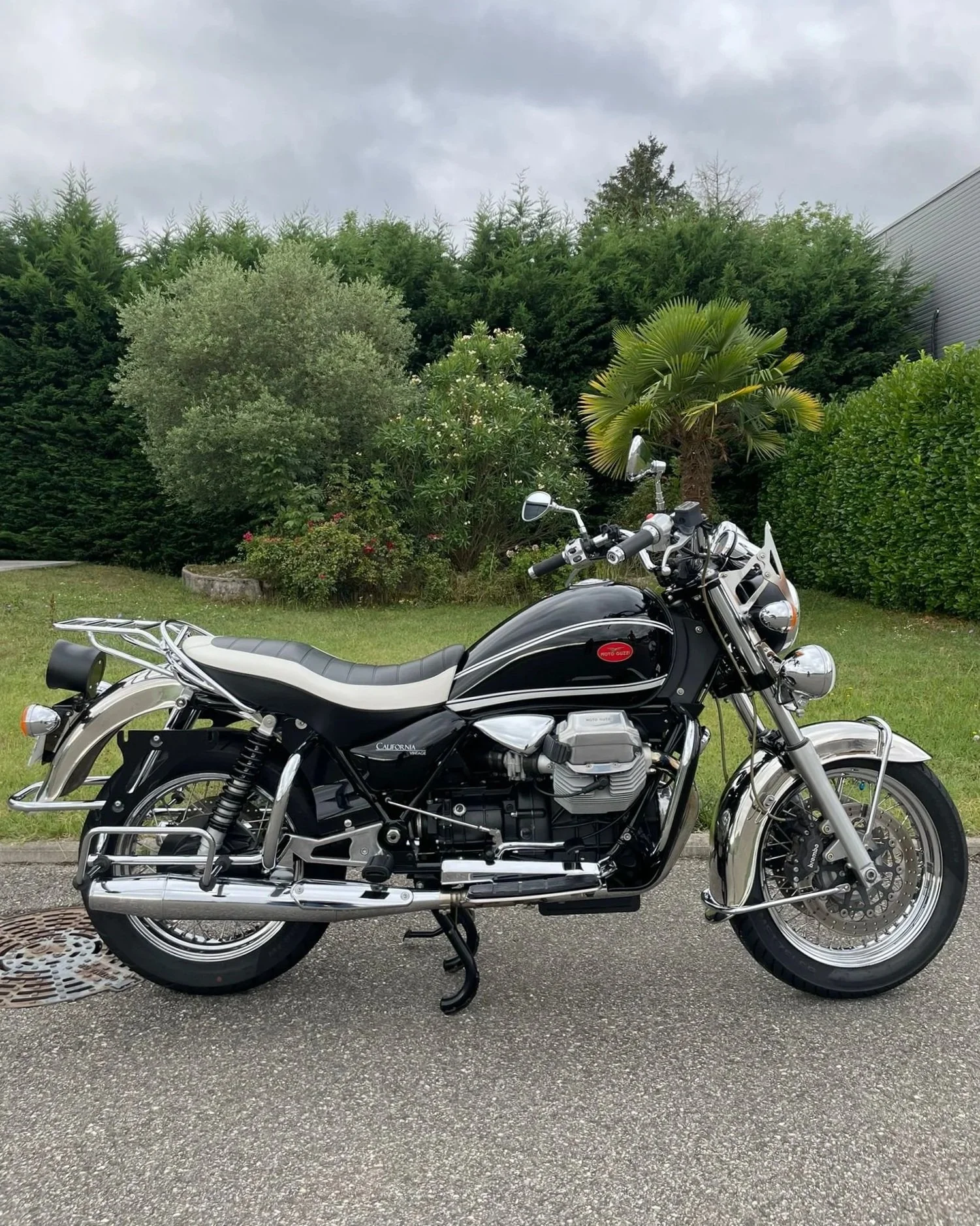 Une moto classique noire et argentée stationnée sur le trottoir devant un jardin avec des arbres et des buissons, sous un ciel nuageux.