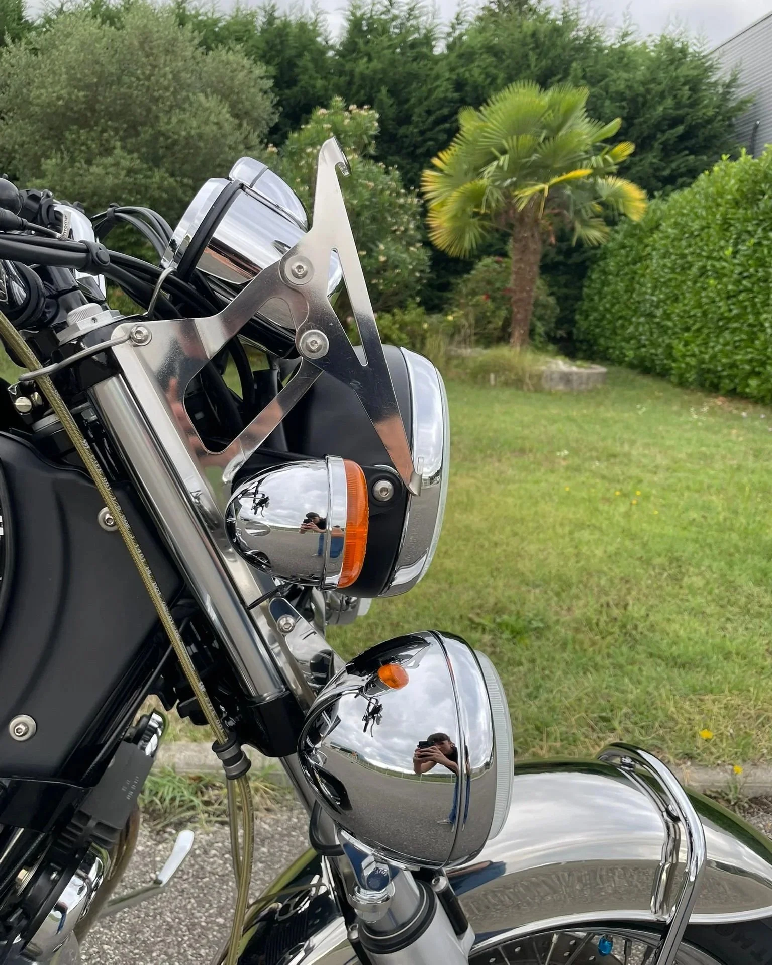 Partie avant d'une moto avec phares chromés, rétroviseurs, et une petite décoration, avec un arrière-plan de jardin contenant des arbres et une pelouse verte.