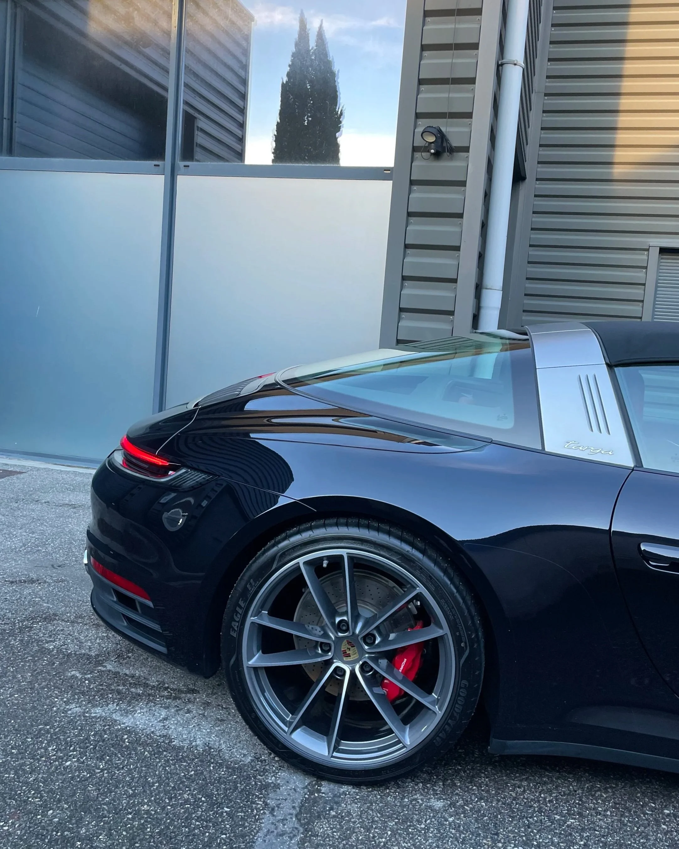 Une voiture noire Porsche cabriolet garée à côté d'un mur industriel en métal, avec un arbre en fond, vue partielle de l'arrière de la voiture, avec un phare arrière et une roue en alliage avec un frein à disque rouge.