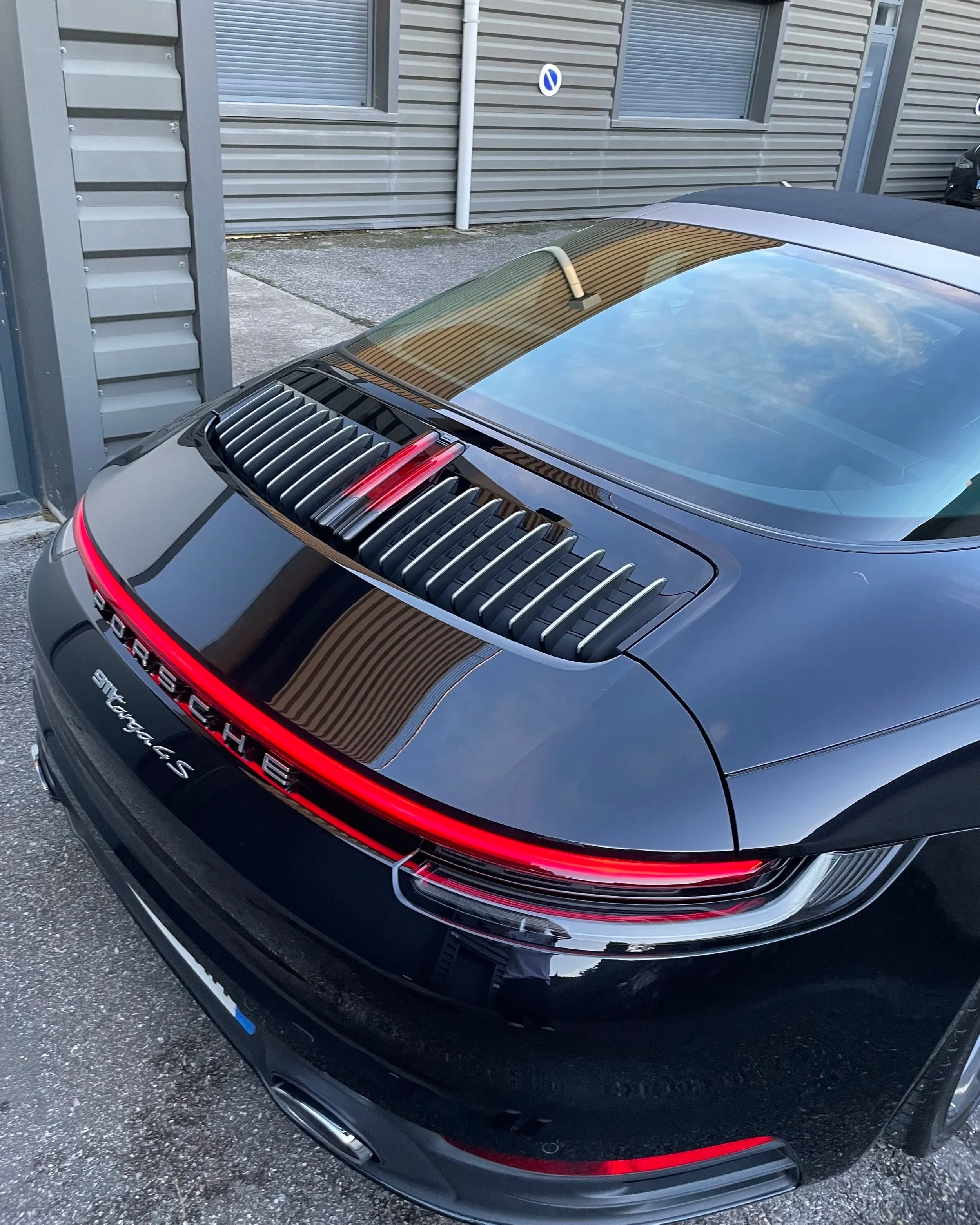 Voiture noire Porsche 911 Carrera 4S vue de l'arrière, stationnée à côté d'un bâtiment en métal avec des fenêtres.