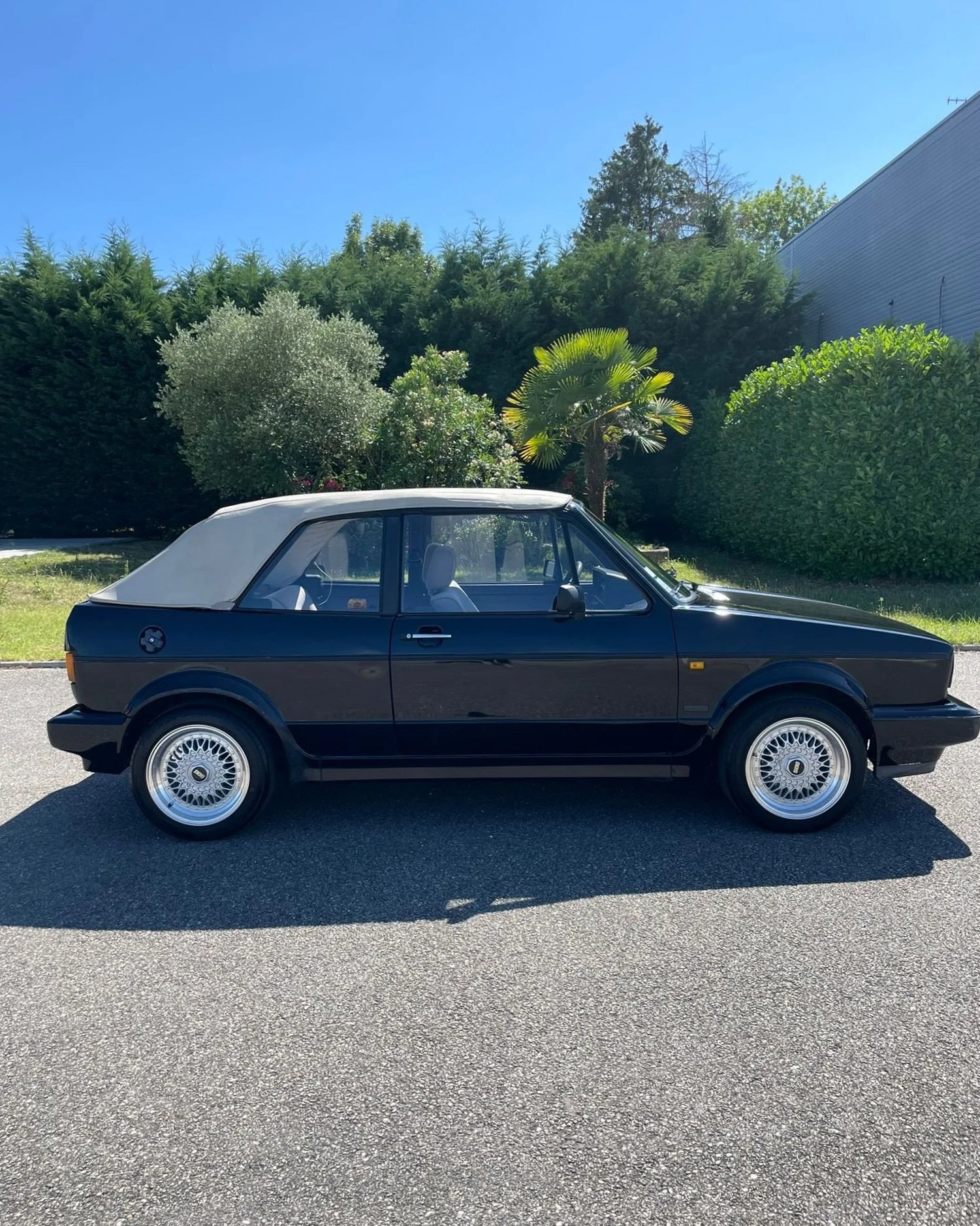Voiture de couleur noire avec toit cabriolet blanc, stationnée sur une route en plein jour, avec des arbres verts en arrière-plan.