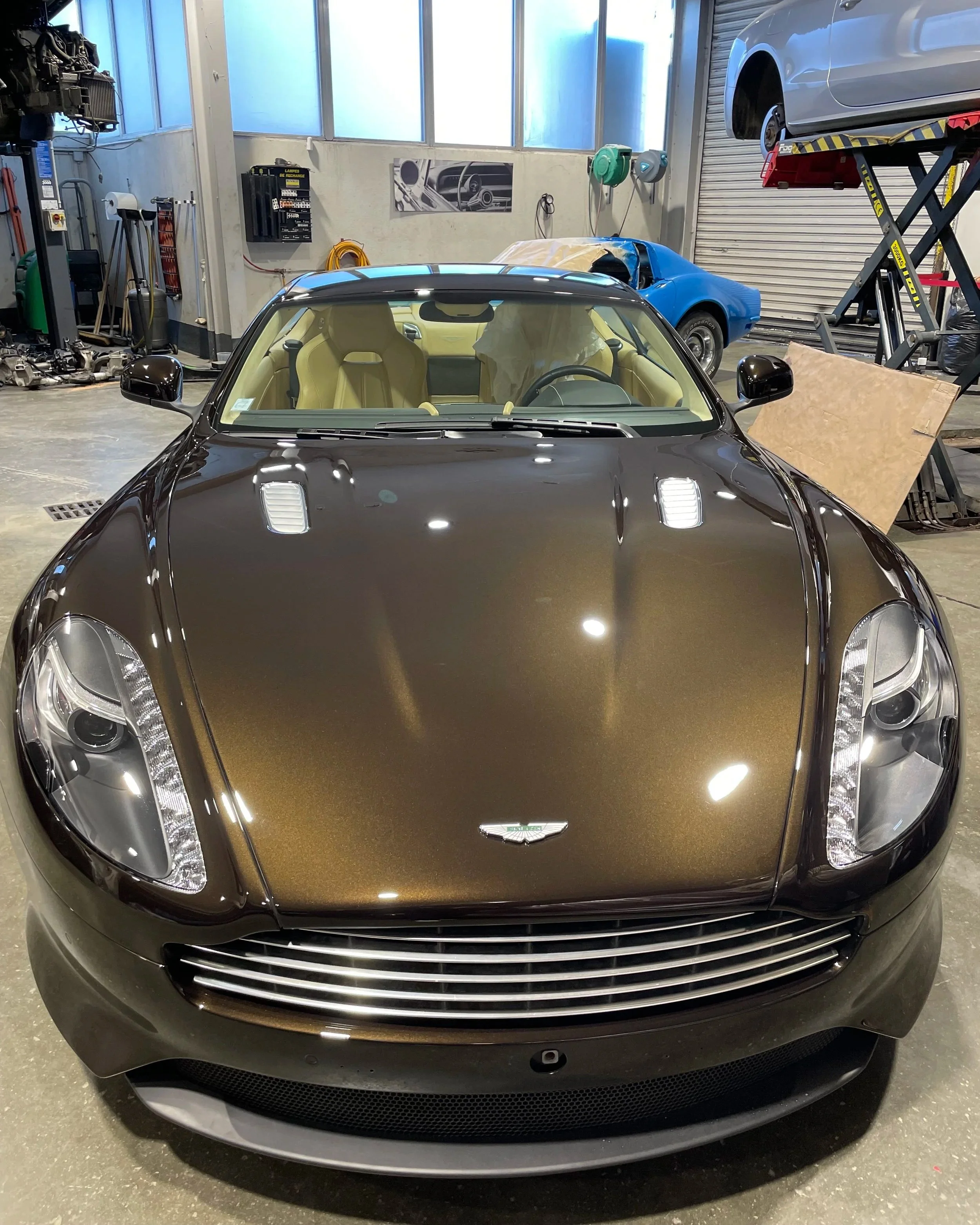 Voiture de luxe de couleur brun foncé dans un garage, vue de face, intérieur beige clair visible à travers le pare-brise, avec deux voitures de sport en arrière-plan.