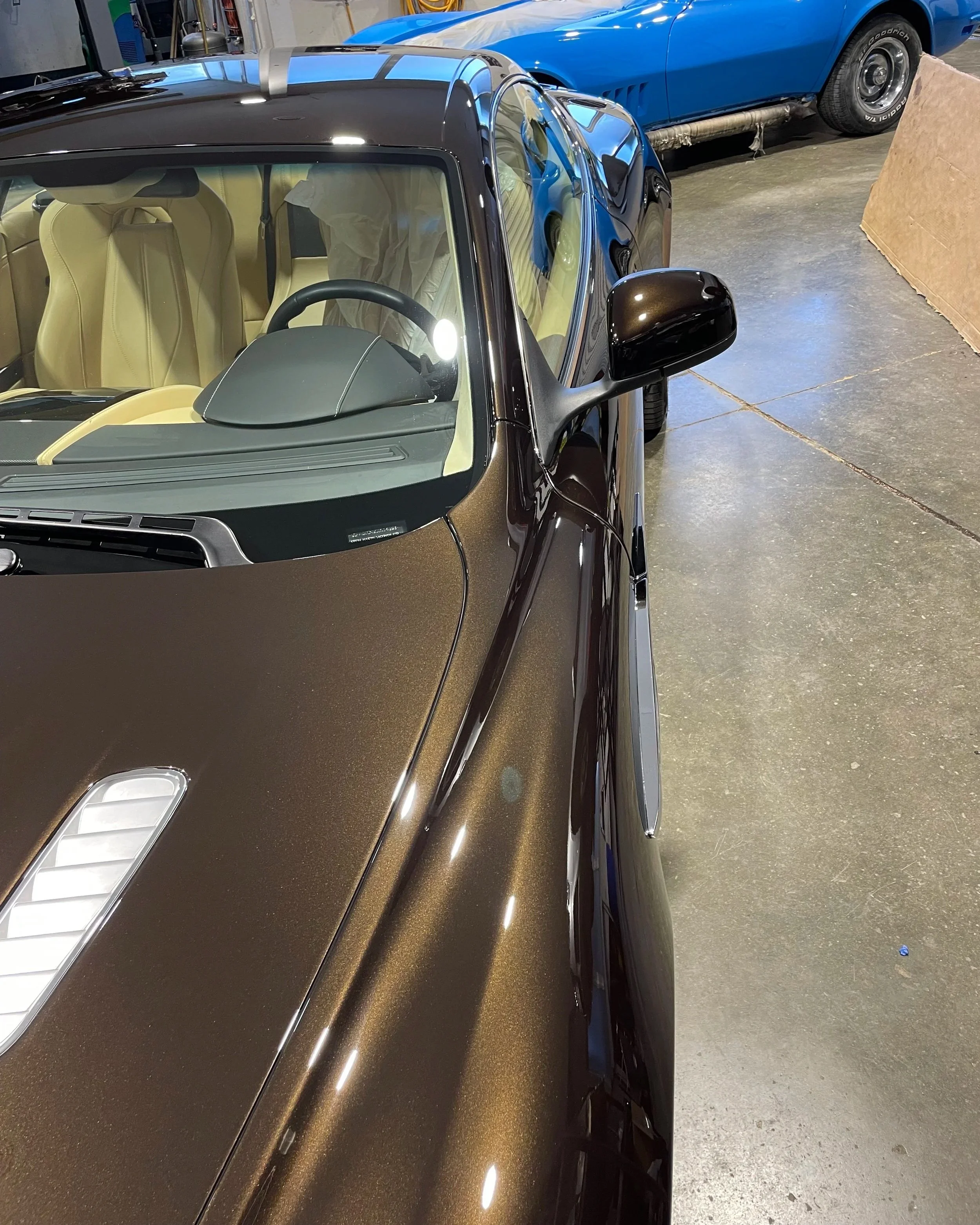 Voiture de sport conçue pour la course, avec teinte marron métallique et intérieur beige clair, stationnée dans un garage, avec une autre voiture bleue en arrière-plan.