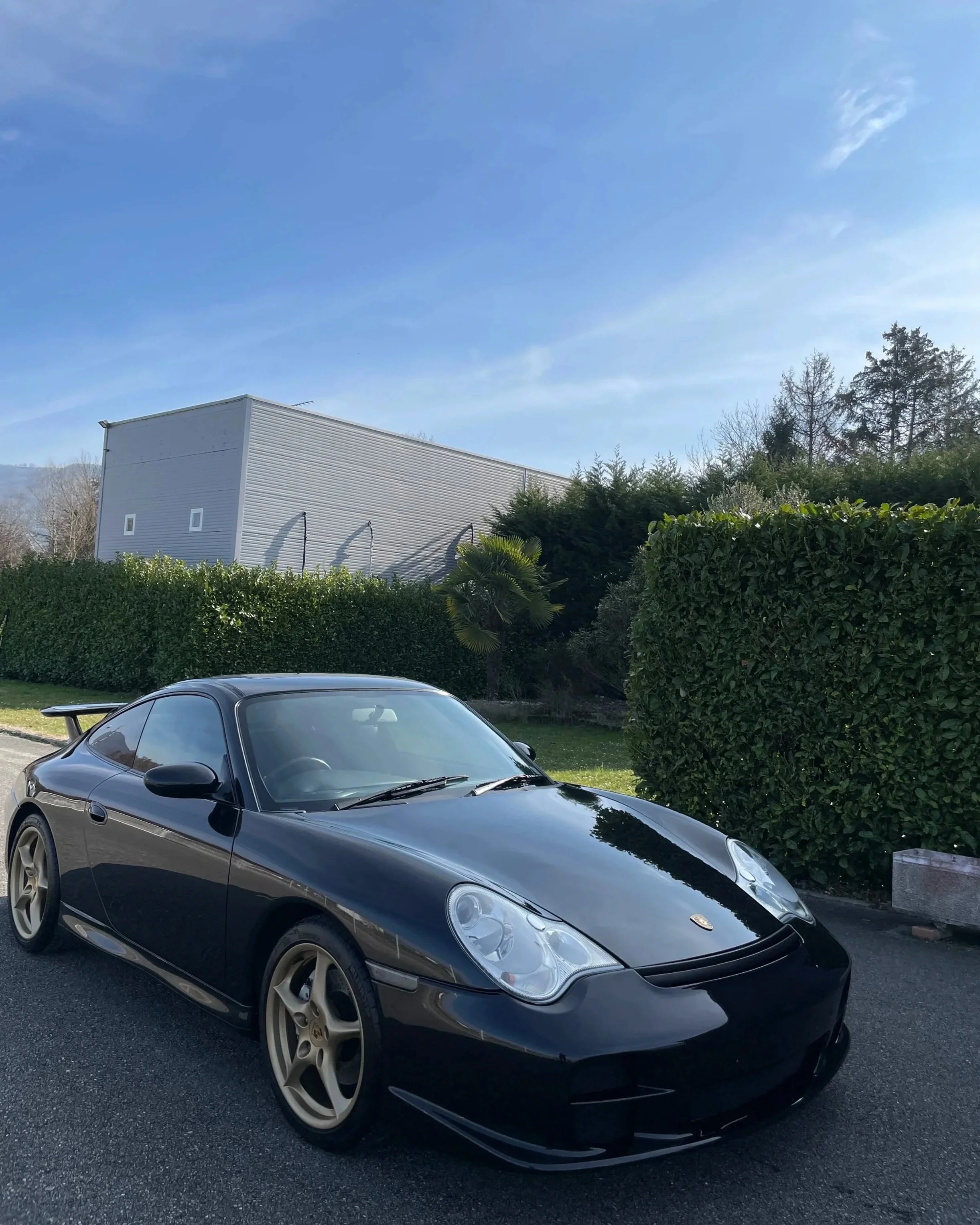 Voiture Porsche Carrera noire stationnée sur le côté d'une route avec des haies vertes et un bâtiment moderne en arrière-plan sous un ciel bleu clair.