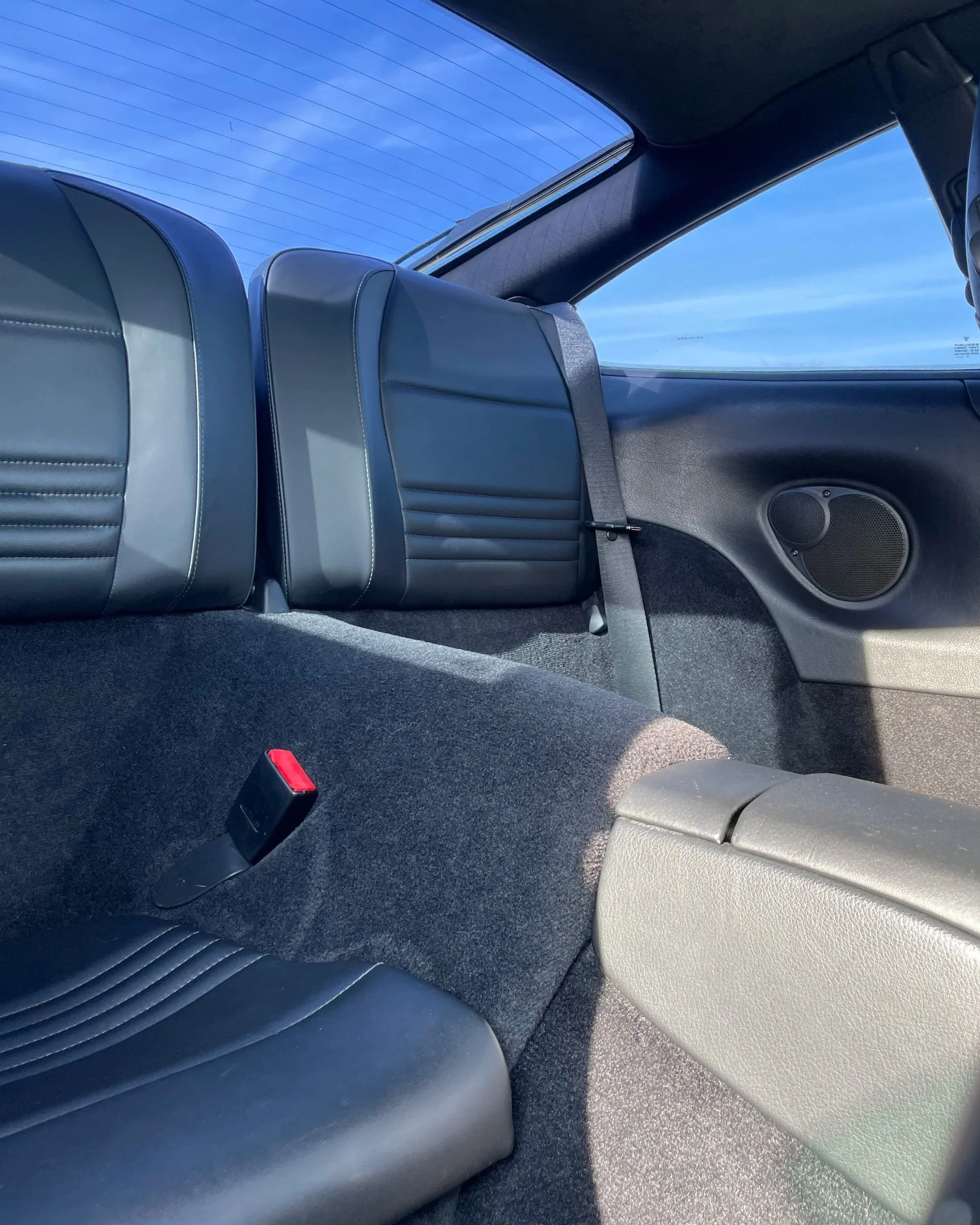 Intérieur d'une voiture, vue de l'arrière avec des sièges en cuir noir, une ceinture de sécurité, une fenêtre arrière avec un ciel bleu visible.