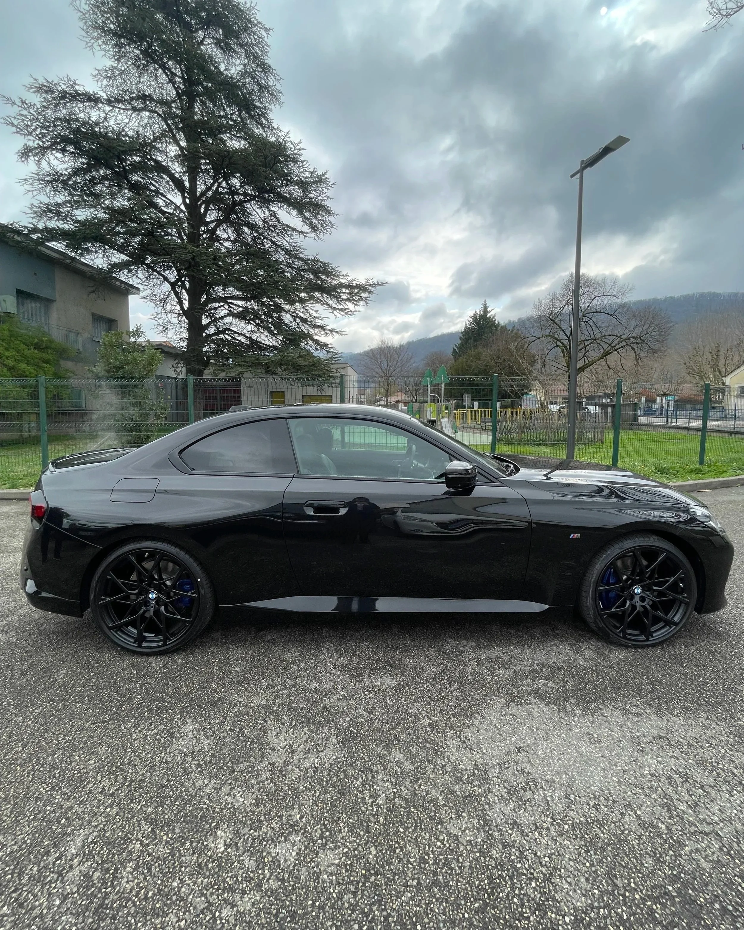 Voiture noire BMW coupé stationnée sur une route, fond avec arbres, clôture, lampadaire et ciel nuageux.