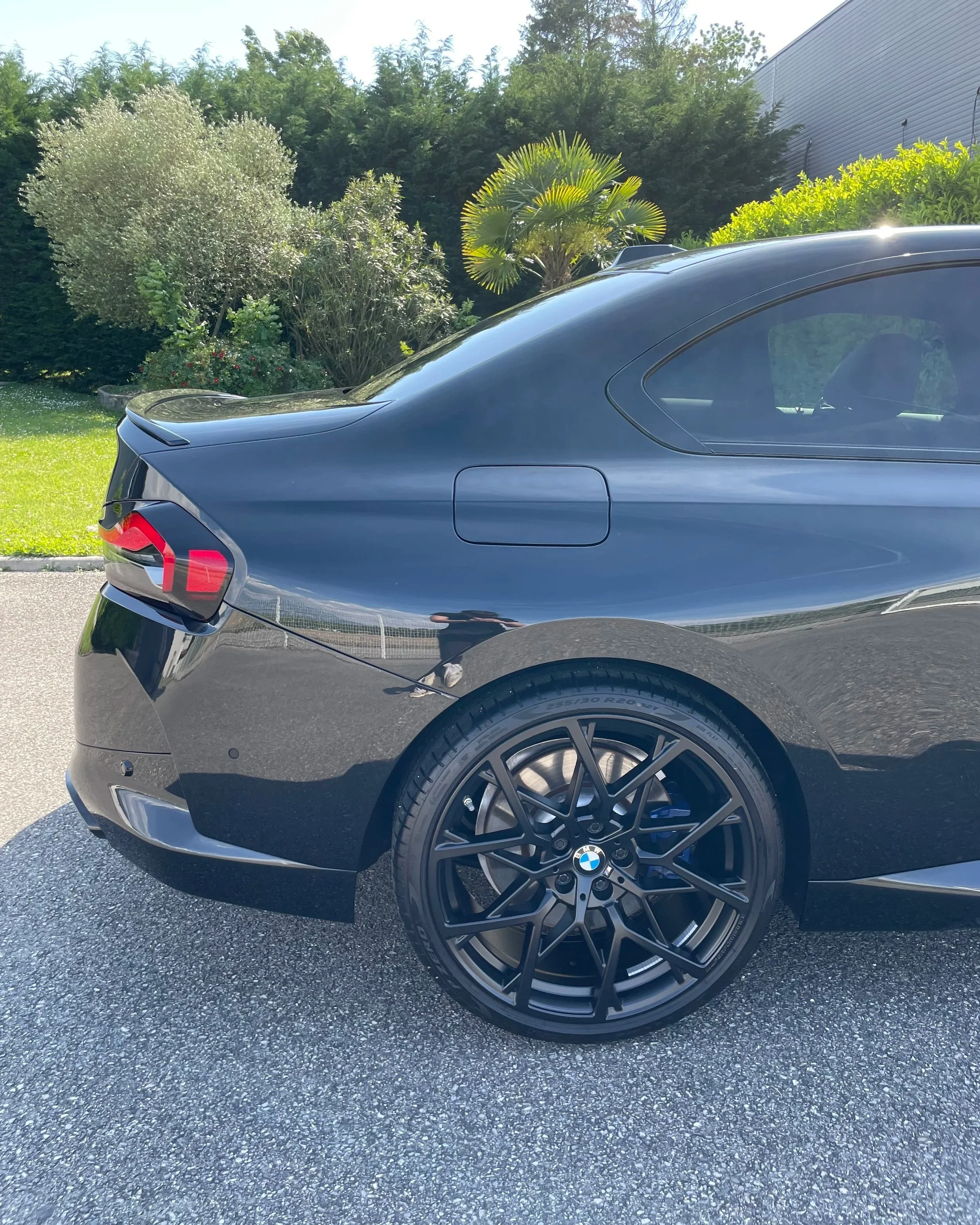 Une voiture noire de marque BMW avec des jantes noires et en arrière-plan, des arbres et un mur en métal