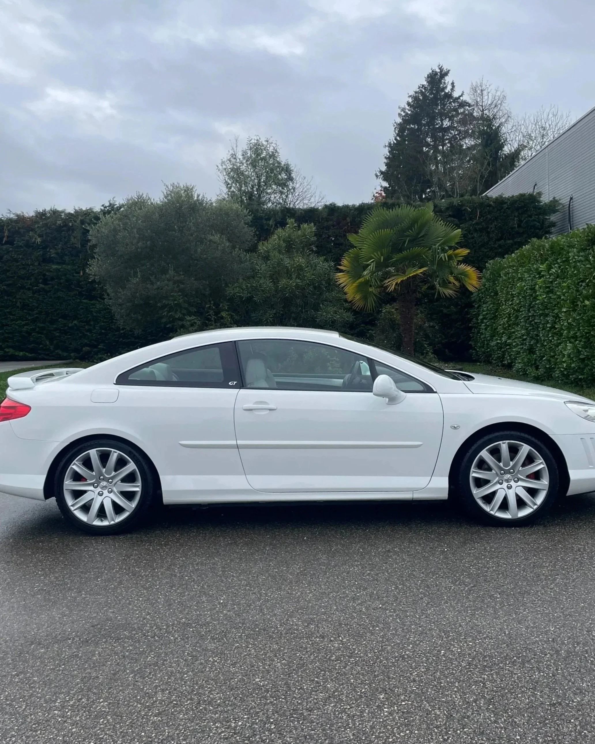 Voiture blanche de marque Mercedes-Benz, modèle coupé, stationnée sur une route pavée avec un fond de végétation comprenant des arbres et un ciel nuageux.